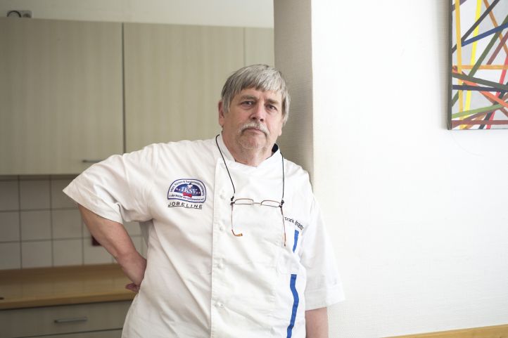 Ein Mann der Koch ist steht in weißer Schürze vor einer Tür und schaut ernst in die Kamera