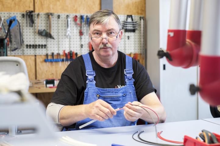 Mann im Blaumann sitzt in einer Werkstatt an einer Werkbank und repariert ein Kabel