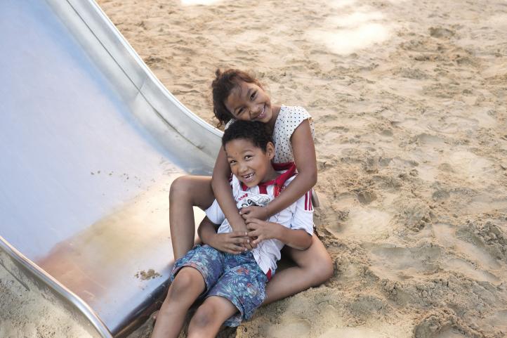 Ein Junge und ein Mädchen sitzen am Fuße einer Rutsche im Sandkasten und umarmen sich lachend