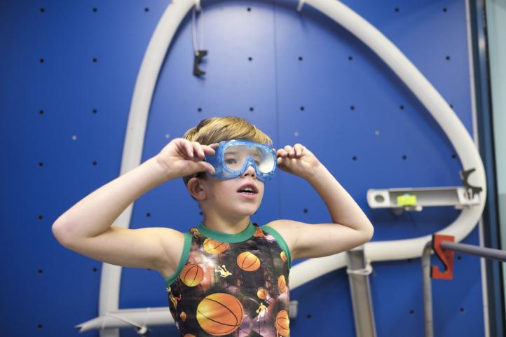 Ein kleiner Junge mit einer Schutzbrille steht in einem Bastelraum vor einer blauen Wand und hat die Hände an die Brille gehoben