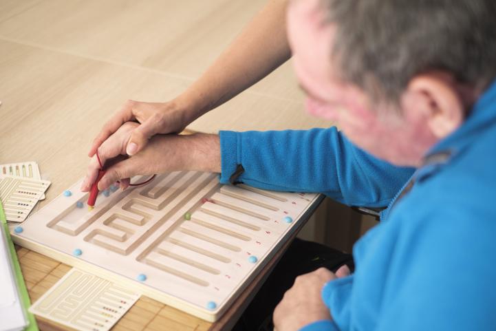 Eine Frauenhand führt die Hand eines Mannes mit einem Holzstift über ein Geschicklichkeitsspiel