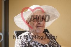 Eine alte Frau Seniorin mit Hut sitzt auf einem Balkon und blickt in die Kamera